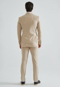 Costume beige avec une coupe ajustée, doté d'une fente unique à l'arrière, et d'un pantalon assorti. Texture lisse et léger éclat. Chaussures noires visibles.