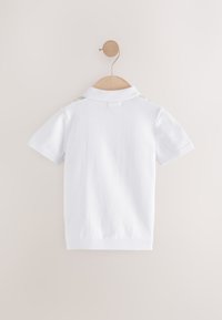 Biela polokošeľa visí na drevenom ramienku, krátke rukávy, textúrovaná látka, rebrovaný golier a viditeľné švy na pleciach.