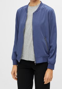 Bomberjack in blauwe stof met een ritssluiting aan de voorkant, geribde manchetten en accentuering met zijzakken, gecombineerd met een lichtgrijs shirt eronder.