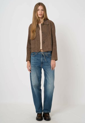 Jonge vrouw met lang haar, gekleed in een bruin jasje met knopen, een beige top, losse blauwe spijkerbroek met een koordriem en donkerbruine schoenen.