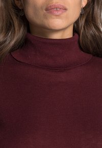 Femme portant des boucles d'oreilles argentées rondes à motif et un pull à col roulé côtelé bordeaux, lèvres légèrement entrouvertes, cheveux détachés.