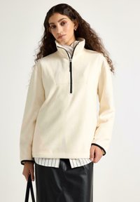 Žena s dlouhými kudrnatými vlasy, oblečená v krémovém fleecovém svetru se černým zipem, černé kožené sukni a pruhované košili, držící černou tašku.