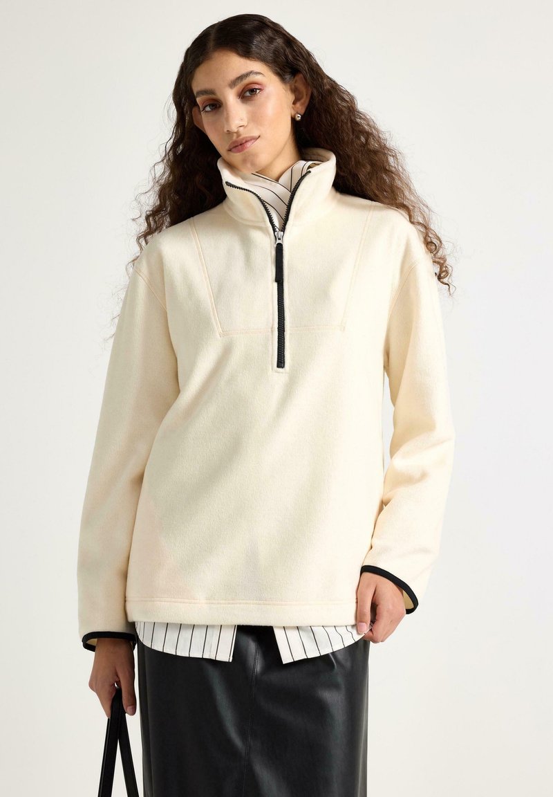 Žena s dlouhými kudrnatými vlasy, oblečená v krémovém fleecovém svetru se černým zipem, černé kožené sukni a pruhované košili, držící černou tašku.