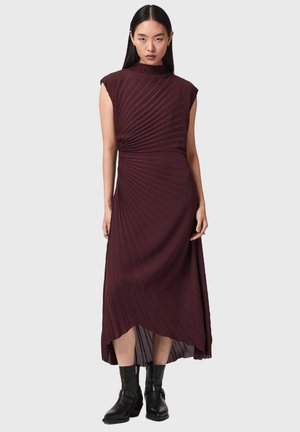 Frau in einem ärmellosen, plissierten dunkelvioletten Midikleid mit asymmetrischem Saum, kombiniert mit schwarzen Stiefeletten, steht vor einem schlichten Hintergrund.