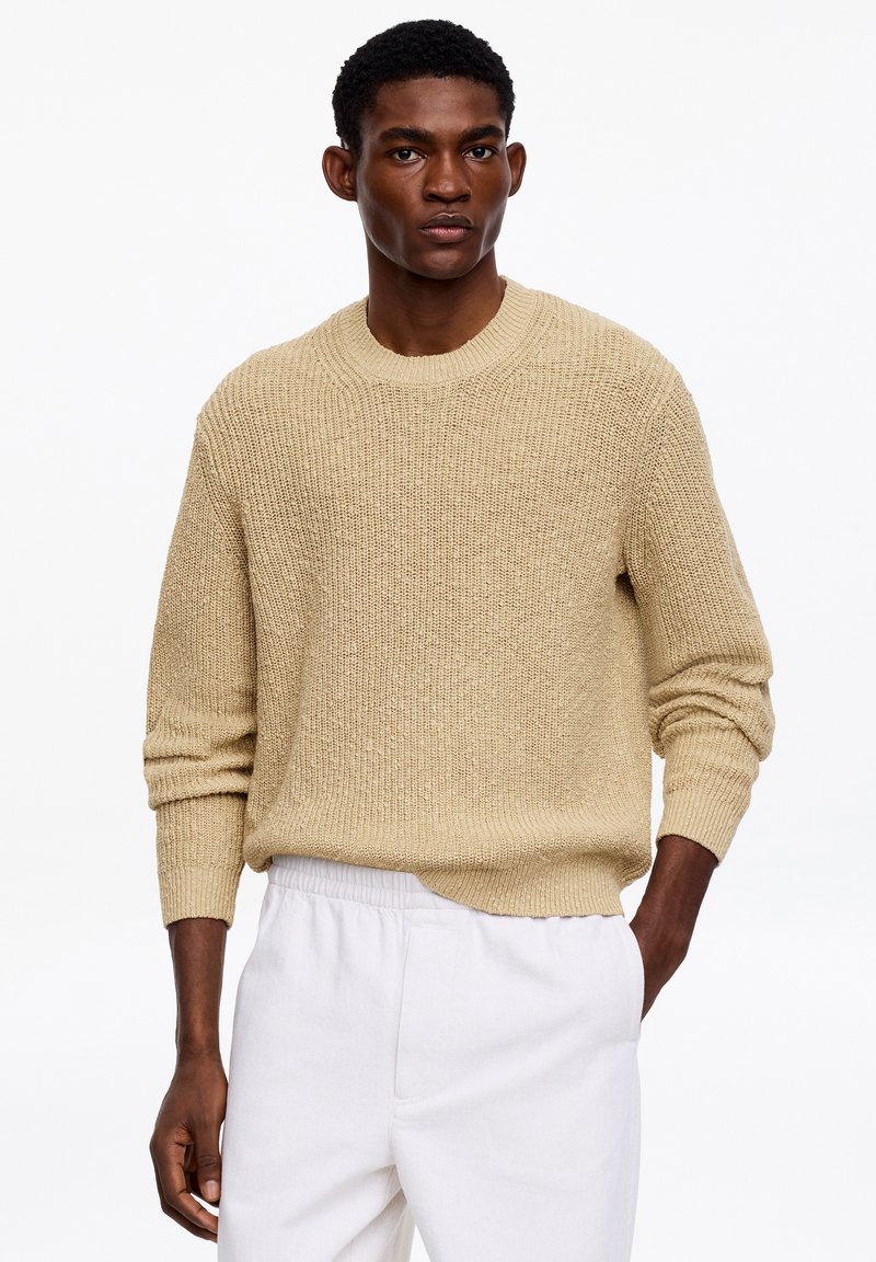 Mann trägt einen beige gestrickten Pullover und weiße Hose, steht mit einer Hand in der Tasche vor einem schlichten weißen Hintergrund.