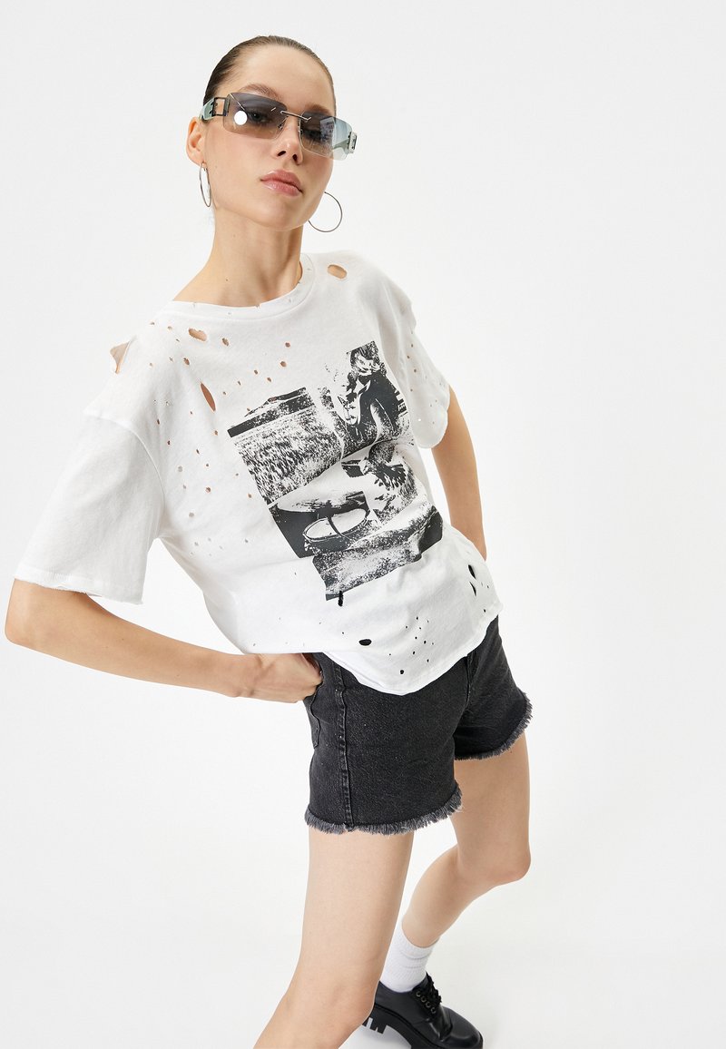 Hvid distressed T-shirt med grafisk print, parret med sorte denimshorts med frynsede kanter. Accessories inkluderer solbriller og hoops øreringe.