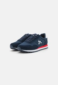 Sneaker blu navy con un mix di materiali lisci e texture, con suola bianca e striscia di accento rossa. Logo sul lato esterno.