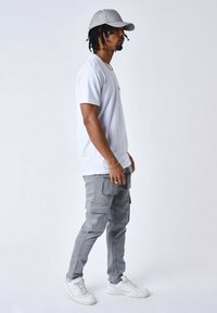 T-shirt bianca, pantaloni cargo grigi con tasche e sneakers bianche. Cappellino grigio a motivo, maniche corte e vestibilità ampia. Materiali testurizzati visibili.