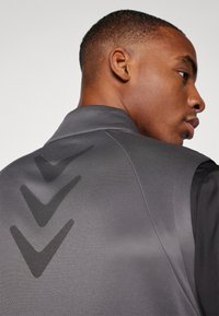 Gilet de sport gris avec une finition texturée, présentant un motif en chevrons noir distinctif dans le dos et un design de col structuré.