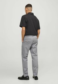 Uomo in piedi con le mani in tasca, che indossa una polo nera a maniche corte, pantaloni grigio chiaro e scarpe nere, rivolto lontano dalla fotocamera.