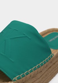 Βερμεγιόν σανδάλι σε τυρκουάζ με ανάγλυφα αρχικά στο φαρδύ λουράκι και πλεγμένη σόλα από τζούτ σε πλατφόρμα, σε λευκό φόντο.