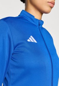 Μπλε αθλητικό μπουφάν με ψηλό γιακά, ολόκληρο φερμουάρ και λευκή λεπτομέρεια λογότυπου. Κατασκευασμένο από υφασμάτινη υφή, ελαφρύ ύφασμα.