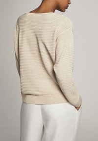 Maglione a righe beige chiaro con un ampio scollo, orlo e polsini a costine. La texture sembra morbida e leggermente strutturata. Abbinato a pantaloni bianchi.