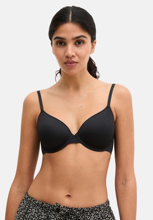 Kobieta o ciemnych włosach spiętych do tyłu, ubrana w czarny biustonosz i czarne kwieciste majtki, zwrócona przodem na jasnym, jednolitym tle.