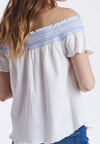 Top bianco scollato con dettagli in smocking blu, tessuto texturizzato, maniche a balze e vestibilità ampia, abbinato a jeans in denim.