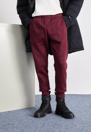Osoba nosząca bordowe spodnie dresowe Nike z czarnymi logo, czarne buty na sznurowadła, białą koszulę i czarną kurtkę, stojąca na niebieskim dywanie.