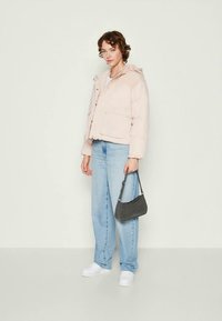 Sieviete, tērpusies gaiši rozā pūkainajā jaku, baltos sporta apavos un gaiši zilos džinsos, tur nelielu melnu rokassomu uz viendabīga balta fona.