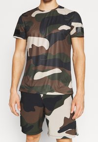 Koszulka sportowa w wzór kamuflażu w ziemistych odcieniach brązu, zieleni i czerni, z krótkimi rękawami oraz okrągłym dekoltem, zaprojektowana z myślą o komforcie.