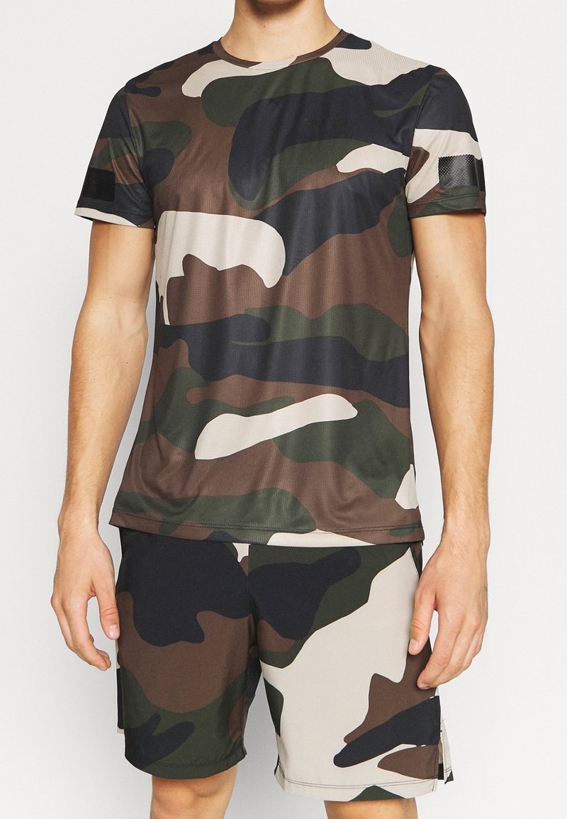 Koszulka sportowa w wzór kamuflażu w ziemistych odcieniach brązu, zieleni i czerni, z krótkimi rękawami oraz okrągłym dekoltem, zaprojektowana z myślą o komforcie.