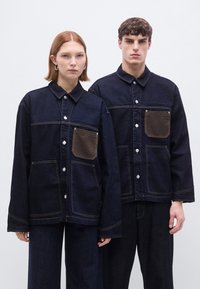 Σκούρα τζιν μπουφάν με αντίθετες καφέ τσέπες, ασημένια κουμπιά και εμφανείς ραφές, σε συνδυασμό με τζιν παντελόνια που ταιριάζουν.