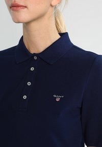 Donna che indossa una polo GANT blu navy con colletto bottone e logo sul petto, sfondo chiaro.