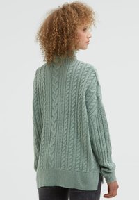 Hellgrüner, grob gestrickter Pullover mit hohem Kragen, übergroßer Passform und geripptem Saumdetail. Die Textur ist weich mit auffälligen Flechtmustern.