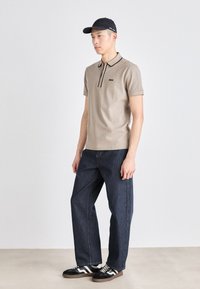 Béžové polo tričko s černými akcenty, volné tmavě modré džíny a černá kšiltovka. Obuv má černobílý pruhovaný design.