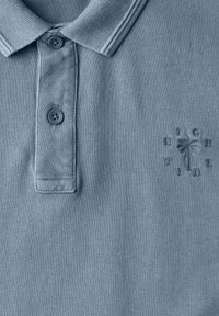 Ljusblå pikétröja, strukturerad tygkvalitet, två knappar, och en broderad palmträd-logotyp med texten "HIGH TIDE" runtomkring.