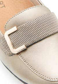 Chaussure à enfiler en métal doré avec une bande élastique texturée et une boucle rectangulaire en ton or, accompagnée de détails de couture contrastants.