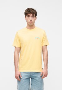 Νέος με κοντά σγουρά μαλλιά που φοράει κίτρινο μπλουζάκι Rip Curl και ανοιχτό μπλε τζιν σορτς, στέκεται μπροστά σε λευκό φόντο.