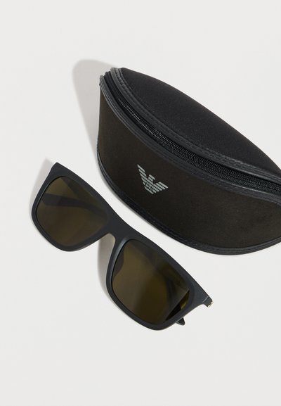 Μαύρα ορθογώνια γυαλιά ηλίου με σκουρόχρωμους φακούς δίπλα σε μαύρη σκληρή θήκη με φερμουάρ που φέρει ασημένιο λογότυπο αετού.