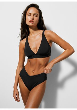 Młoda kobieta w czarnym dwuczęściowym stroju kąpielowym z głębokim dekoltem w kształcie litery V i majtkami z wysokim stanem, stojąca na jasnym, jednolitym tle.