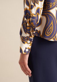 Blusa a maniche lunghe con base bianca e audaci motivi floreali blu navy e oro. Include polsini con bottoni e una texture di tessuto liscia.
