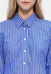 Blonde Person in einem blauen Hemd mit weißen vertikalen Streifen und weißen Knöpfen, Kragen am oberen Ende geknöpft.
