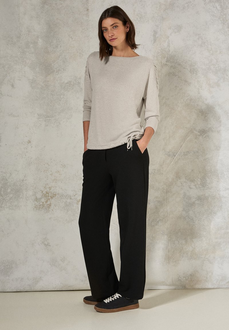 Maglietta grigia chiara a maniche lunghe con dettaglio arricciato sulle spalle, abbinata a pantaloni neri a gamba larga e sneaker scure con lacci bianchi.