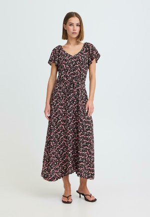 Kvinde iført en sort midi-kjole med pink blomsterprint, korte flagermusærmer, V-hals og sorte sandaletter med hæl, stående på en ensfarvet baggrund.