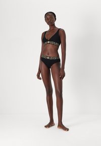 Melna bikini komplekts ar zelta grieķu atslēgas rakstu, kas sastāv no braletes augšdaļas un zemu augšdaļu biksēm, izgatavots no gludas auduma, minimālistisks dizains.