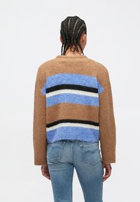 Pulover maro tricotat cu un design cu dungi în bloc în albastru, negru și alb. Are o croială lejeră și mâneci lungi, asortat cu blugi albaștri.