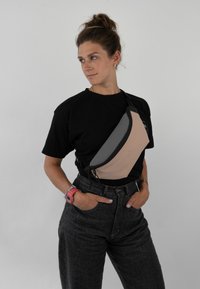 Beige és szürke színű sling táska, sima anyagból, ívelt cipzárral, a mellkas felett viselve. Fekete póló és sötét farmer látható.