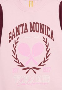 Ružová mikina s marhuľovými rukávmi. Obsahuje text "SANTA MONICA Lawn Club" a "Tennis Club", grafiku veniec a dizajn prekrížených rakiet.