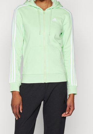 Persoon draagt een lichtgroene Adidas hoodie met rits met witte strepen op de mouwen en zwarte Adidas broek, staande tegen een effen achtergrond.