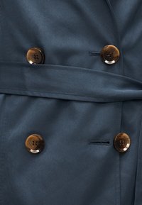 Marineblauwe stof met vier ronde bruine knopen, gerangschikt in paren, en een bijpassende stoffen riem over het middelste gedeelte.