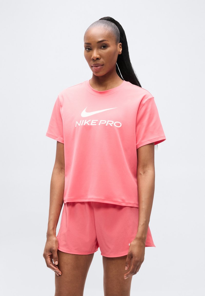 Ροζ αθλητικό τοπ Nike Pro με κοντά μανίκια, με λευκό λογότυπο. Συνδυάζεται με ροζ σορτς ίδιας απόχρωσης. Λείο, ελαφρύ ύφασμα.