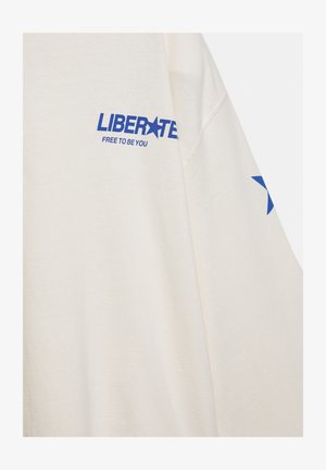 Cămașă cu mânecă lungă de culoare crem, cu text albastru „LIBERATE FREE TO BE YOU” și o stea albastră pe mânecă.
