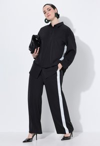Schwarzes Hemd mit Knöpfen und lässigem Schnitt, kombiniert mit schwarzen Hosen, die einen seitlichen weißen Streifen haben; das Modell hält eine schwarze Clutch und trägt stylische High Heels.