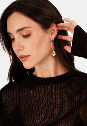 Zlaté náušnice ve tvaru kruhu s zaobleným, kulovitým akcentem. Modelka má na sobě průhledný černý pletený top, s detailním záběrem na náušnice.