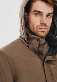 Brązkowa wodoodporna kurtka z kapturem, z zamkiem błyskawicznym, teksturowanym materiałem i szarym polarem w środku. Zbliżenie na górną część ciała.