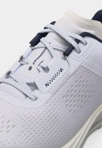 Κοντινό πλάνο λευκού αθλητικού παπουτσιού με υφασμάτινη υφή, ανοιχτό γκρι κορδόνια, ναυτικού χρώματος τρύπες και την επιγραφή «HOVR AVENGER 2» στη γλώσσα.