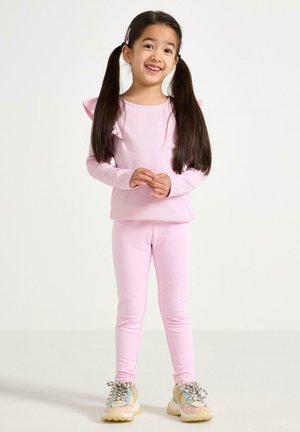 Lyserød langærmet top med flæse detaljer ved skuldrene, kombineret med matchende lyserøde leggings og flerfarvede sneakers. Overordnet glat tekstur.