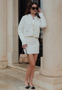 Weißes strukturiertes Blazer- und Bleistiftrock-Set, kombiniert mit einer beigefarbenen Handtasche und schwarz-weißen flachen Schuhen. Zubehör umfasst Sonnenbrillen.